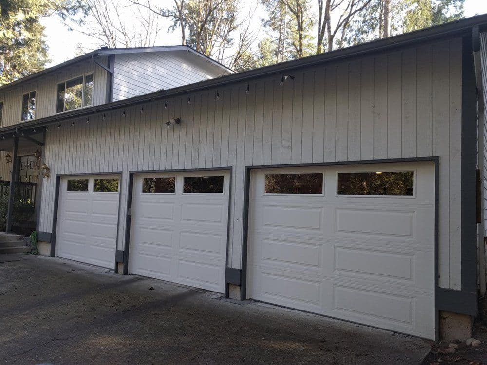 Modern house exterior with three white garage doors and wooded background.