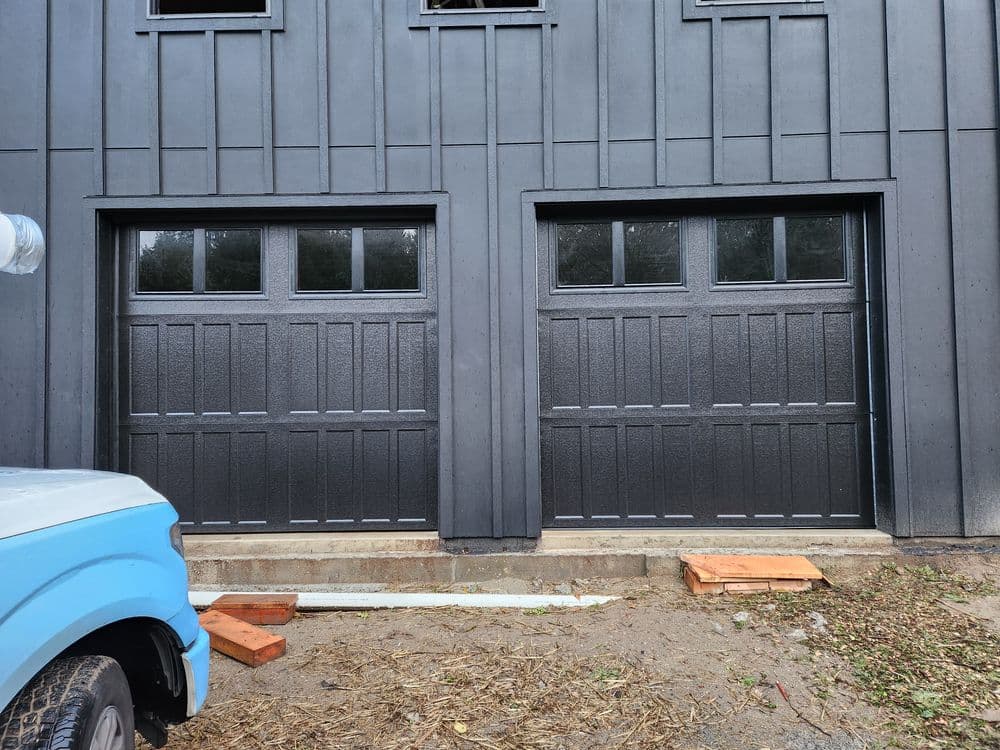 Black wooden garage doors with windows, situated below a gray building exterior.